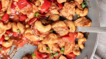 A spoonful of stir-fried chicken, red bell peppers, cashews, and green onions, held above a pan of the same dish.