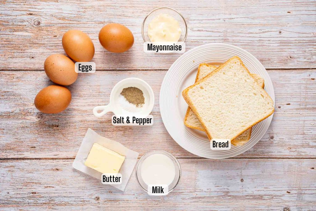 A flat lay of eggs, mayonnaise, sliced bread, butter, milk, and a salt and pepper mix—each labeled—on a wooden surface, showcasing the essential ingredients for a classic Japanese egg sandwich.
