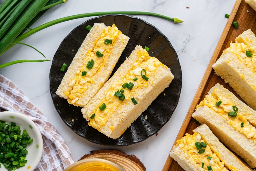 Three Japanese Egg Sandwiches topped with chopped green onions are arranged on a black plate, with more sandwiches and green onions visible nearby.