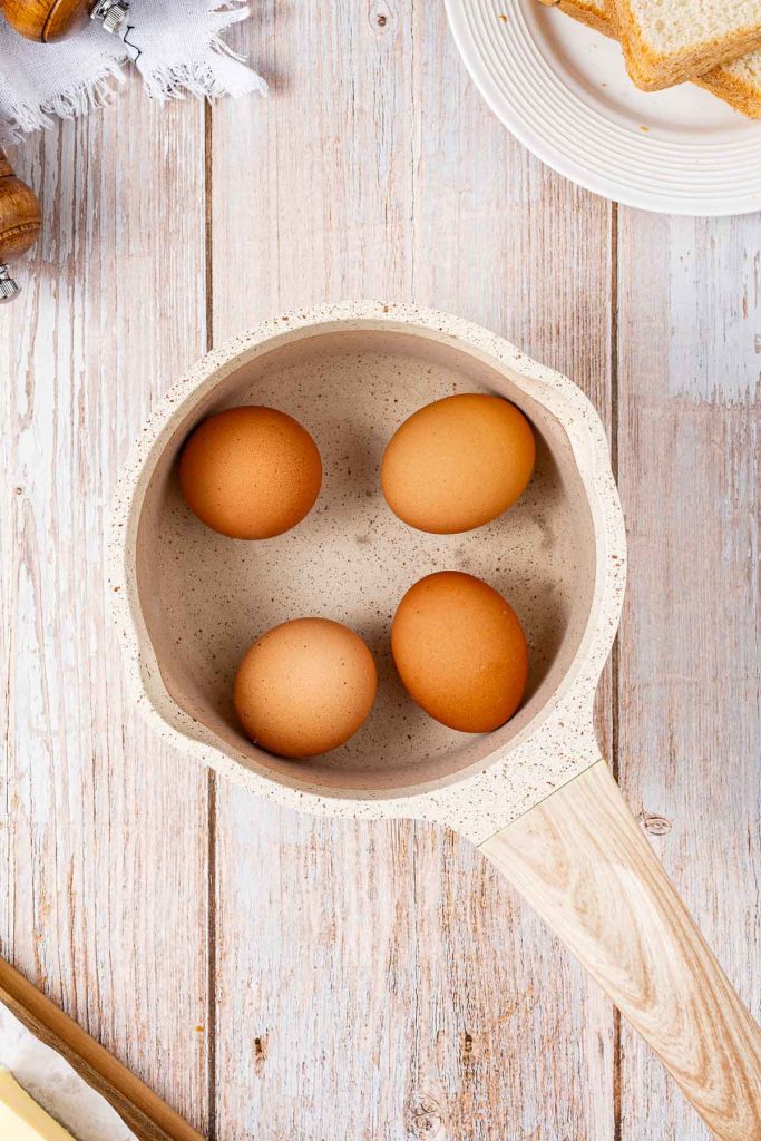 A top-down view of four brown eggs in a cream-colored saucepan on a light wooden surface, with a plate of bread slices nearby—perfect for making a classic Egg Salad Sandwich or Japanese Tamago Sando.