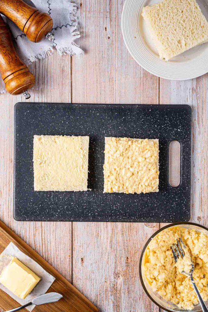 Two slices of bread with egg salad on one slice and butter on the other, placed on a black cutting board with a bowl of Japanese Egg Sandwich filling, butter, and pepper mills nearby.