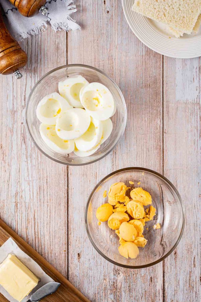 Two bowls on a wooden surface, one with halved hard-boiled egg whites and the other with yolks—classic ingredients for a Japanese Egg Sandwich—alongside bread, butter, and pepper mills.