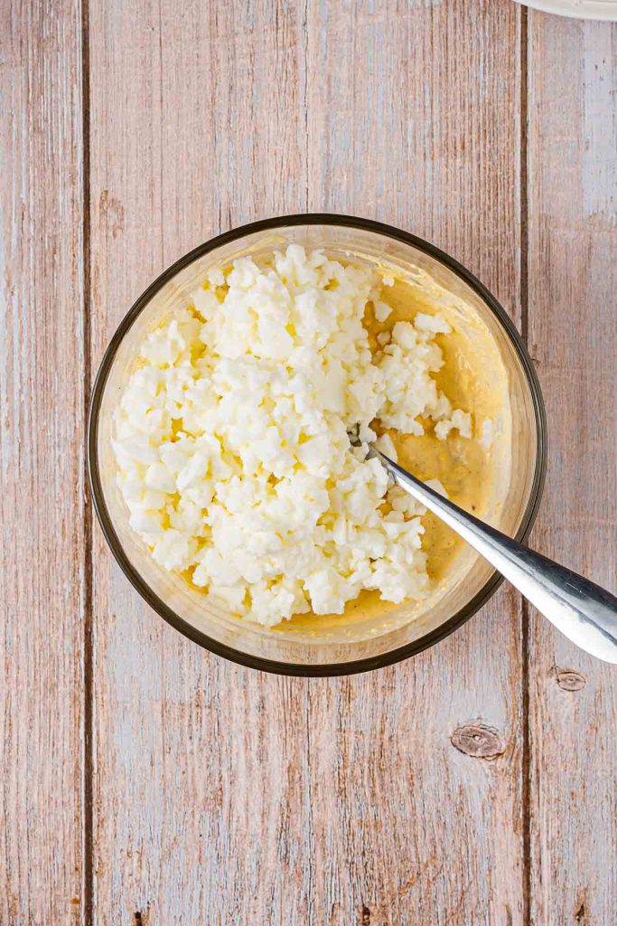 A glass bowl containing a mixture for Japanese Egg Sandwich—with cottage cheese and beaten eggs—sits on a wooden surface with a metal spoon inside, ready to become the perfect filling for Tamago Sando.