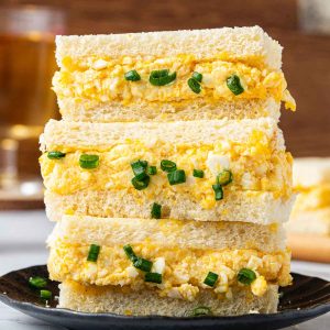 Three Japanese Egg Sandwiches with chopped green onions are stacked on a black plate, featuring soft white bread and a creamy yellow filling.
